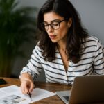 Mulher usando óculos analisa documentos fiscais sobre a mesa enquanto trabalha no notebook, simbolizando o planejamento para regularizar dívidas no PGDAU 11/2025