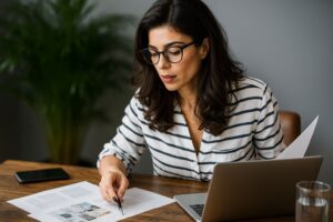 Mulher usando óculos analisa documentos fiscais sobre a mesa enquanto trabalha no notebook, simbolizando o planejamento para regularizar dívidas no PGDAU 11/2025
